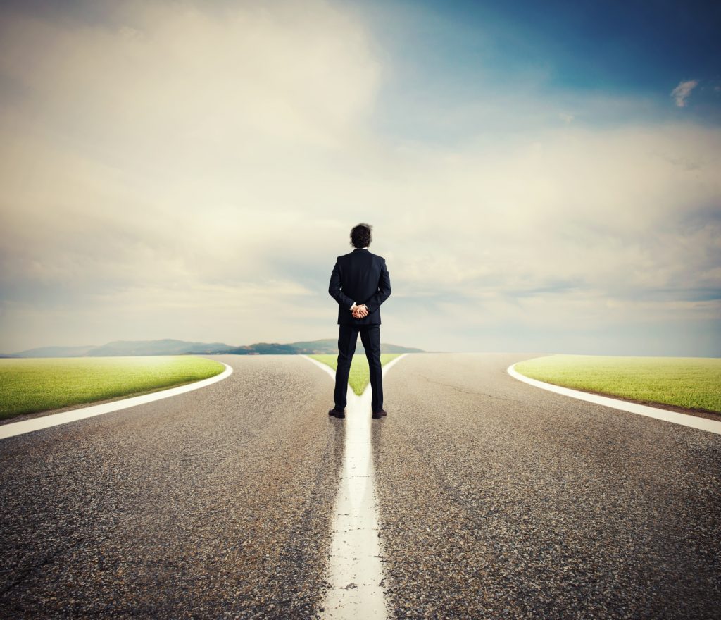 A man in a business suit stands at a crossroads, with one road curving to the left and the other to the right, symbolizing a metaphor for decision-making and choice between floor sealers. The man's back is to the viewer, hands clasped behind him as he contemplates the diverging paths ahead under a dramatic sky.