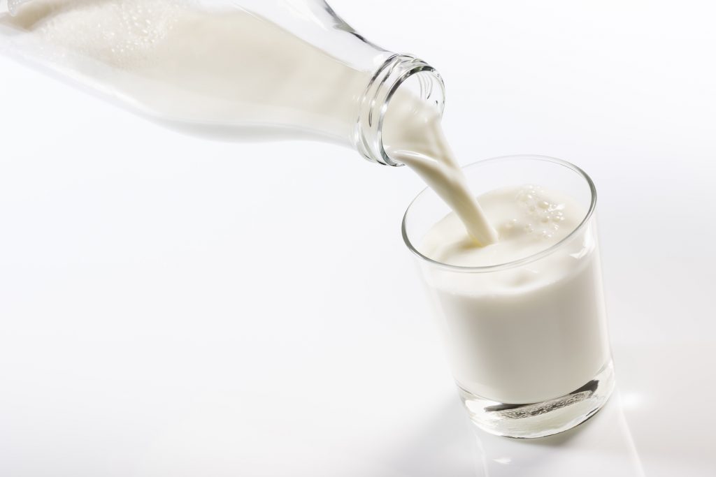 Fresh milk being poured from a clear glass bottle into a transparent glass on a white background. The stream of milk creates a splash and bubbles as it hits the already present milk in the glass, conveying the sense of freshness and purity.
