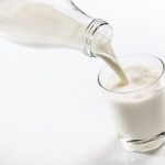 Fresh milk being poured from a clear glass bottle into a transparent glass on a white background. The stream of milk creates a splash and bubbles as it hits the already present milk in the glass, conveying the sense of freshness and purity.