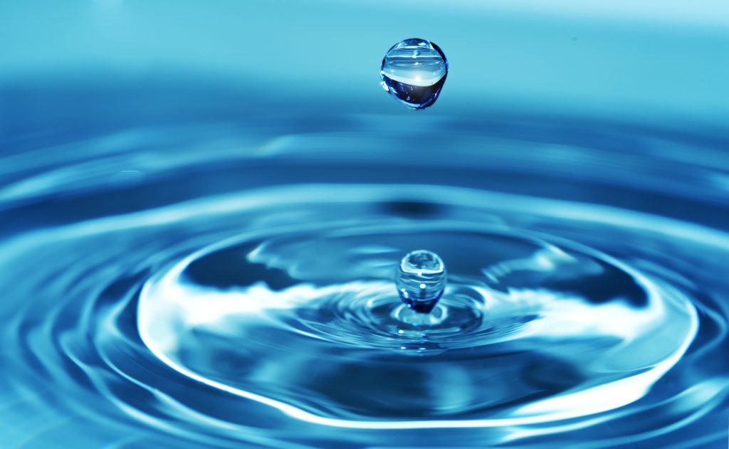 A close-up of a single crystal-clear water droplet suspended above a body of water, creating a series of ripples upon impact. The image captures the droplet in mid-air, with a perfect reflection and the tranquil blue hue of the water providing a serene background.
