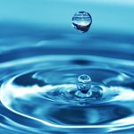 A close-up of a single crystal-clear water droplet suspended above a body of water, creating a series of ripples upon impact. The image captures the droplet in mid-air, with a perfect reflection and the tranquil blue hue of the water providing a serene background.