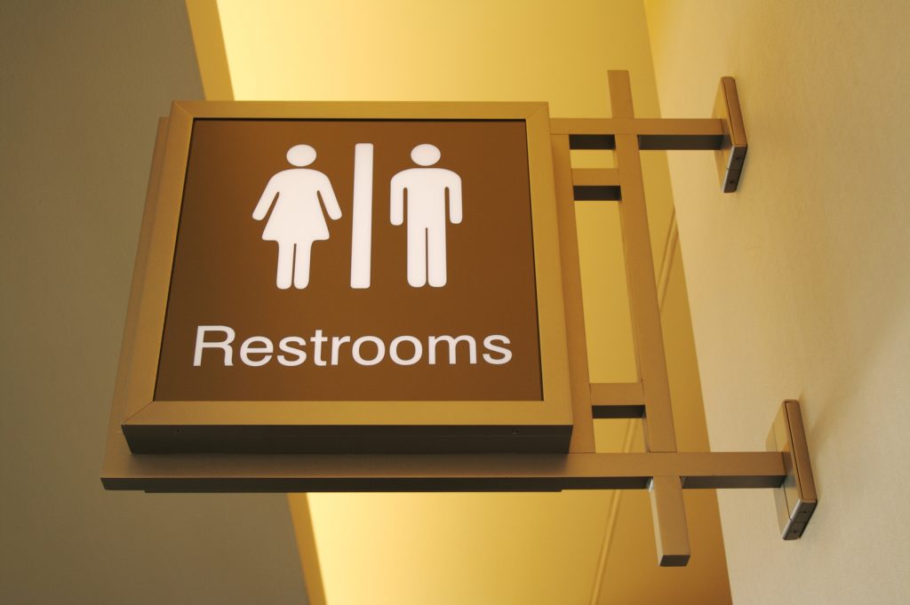 Illuminated restroom sign mounted on a wall, featuring iconography for both male and female facilities. The sign has a warm yellow backlight and is designed with a brown background and white gender symbols, clearly indicating the location of restrooms in a public or commercial setting.