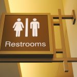 Illuminated restroom sign mounted on a wall, featuring iconography for both male and female facilities. The sign has a warm yellow backlight and is designed with a brown background and white gender symbols, clearly indicating the location of restrooms in a public or commercial setting.