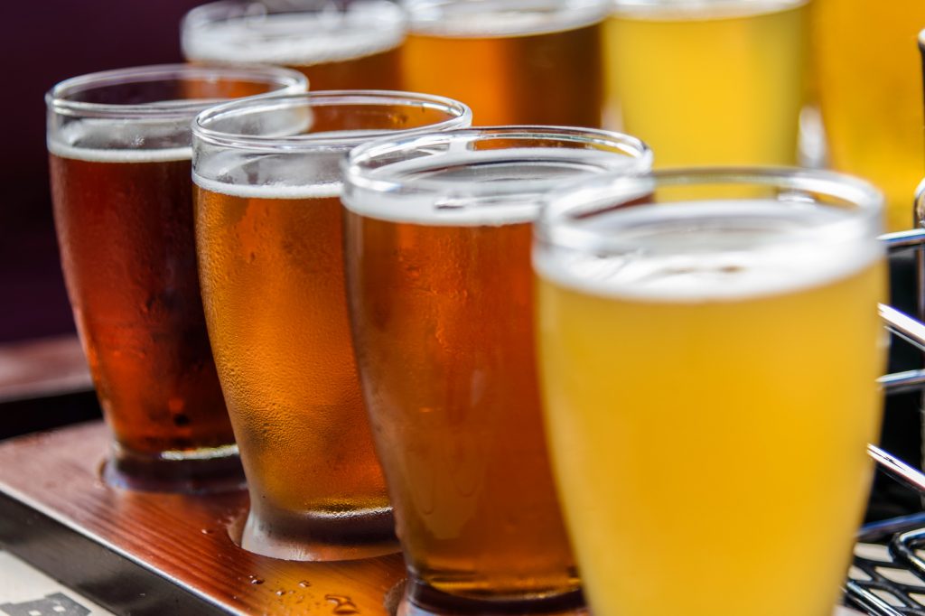 An array of six full beer glasses with varying shades of amber to golden hues, lined up on a dark wooden surface. The glasses reflect light, showcasing the effervescence and clarity of the beers, with the closest glass in sharp focus and the others softly blurred in the background, creating a sense of depth and variety.