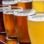 An array of six full beer glasses with varying shades of amber to golden hues, lined up on a dark wooden surface. The glasses reflect light, showcasing the effervescence and clarity of the beers, with the closest glass in sharp focus and the others softly blurred in the background, creating a sense of depth and variety.