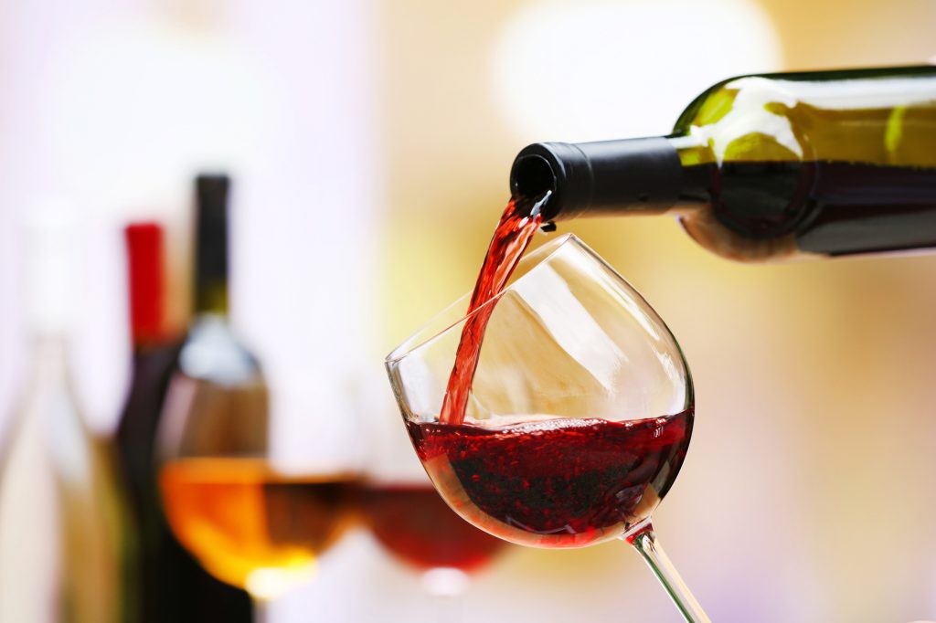 A close-up image capturing the vibrant stream of red wine being poured into a tilted clear wine glass. The background softly blurs a selection of wine bottles, emphasizing the action and the rich color of the wine, with bubbles visible near the surface, suggesting a fresh pour.