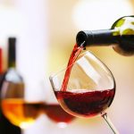 A close-up image capturing the vibrant stream of red wine being poured into a tilted clear wine glass. The background softly blurs a selection of wine bottles, emphasizing the action and the rich color of the wine, with bubbles visible near the surface, suggesting a fresh pour.