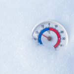 A round outdoor thermometer partially embedded in fresh snow, with the needle indicating a temperature around the freezing point. The thermometer has a clear, legible scale with red and blue color coding for temperature degrees in Celsius, providing a visual contrast against the white snowy background.