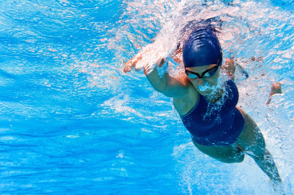 Underwater action shot of a female swimmer mid-stroke in a pool containing chlorine or chlorides, her body angled forward and arm extended. She wears a dark swimsuit, swim cap, and goggles, creating ripples and bubbles in the clear blue water around her, capturing the dynamism and focus of competitive swimming.