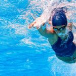 Underwater action shot of a female swimmer mid-stroke in a pool containing chlorine or chlorides, her body angled forward and arm extended. She wears a dark swimsuit, swim cap, and goggles, creating ripples and bubbles in the clear blue water around her, capturing the dynamism and focus of competitive swimming.