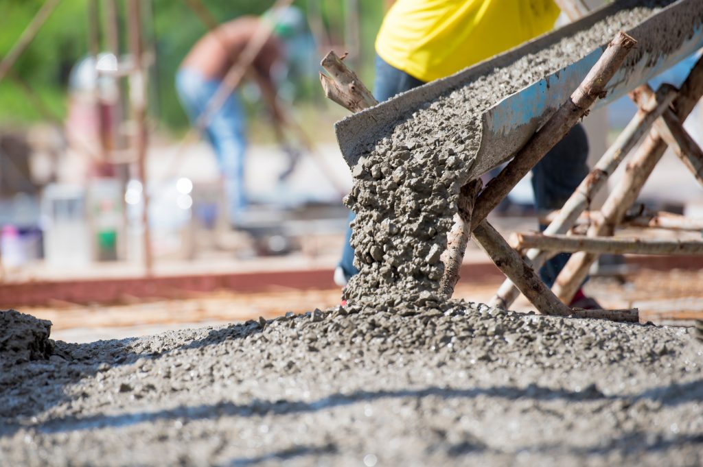 A detailed image of wet concrete being poured from a wheelbarrow onto a construction site, with workers in the background actively involved in the process. The concrete is heaping and spreading, with its rough texture visible, indicative of ongoing construction and building foundations using cement.