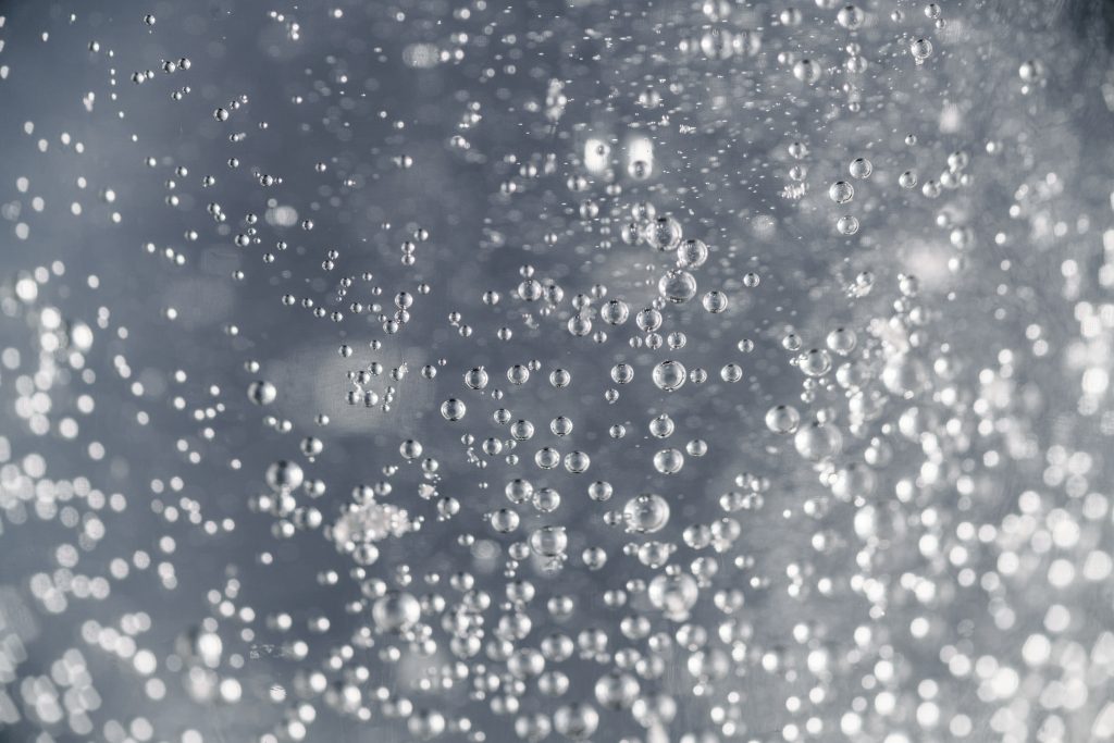 A multitude of sparkling water droplets suspended in liquid due to carbonation, with a focus on their spherical shapes and the way they catch the light. The background is a blurred grey, highlighting the clarity and detail of the individual droplets in this dynamic and refreshing scene.