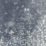 A multitude of sparkling water droplets suspended in liquid due to carbonation, with a focus on their spherical shapes and the way they catch the light. The background is a blurred grey, highlighting the clarity and detail of the individual droplets in this dynamic and refreshing scene.