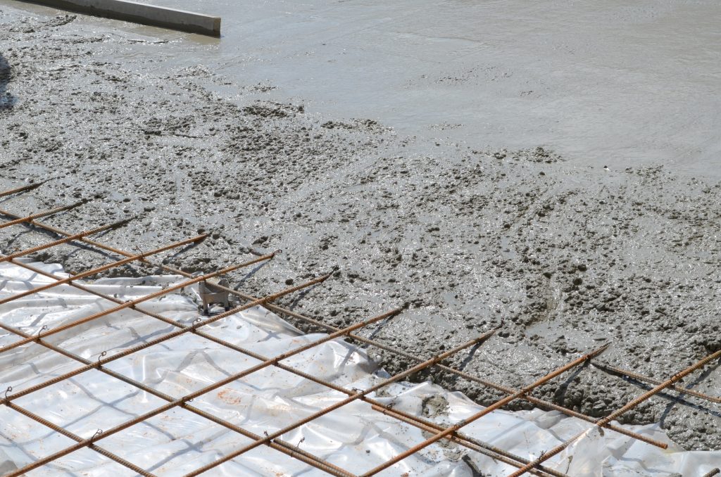 Freshly poured wet concrete over a steel rebar grid foundation, with a section of the grid still exposed. The image captures the process of laying a concrete slab, a crucial step in construction, with the concrete's surface smooth in some areas and roughly textured in others where it has just been spread.