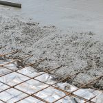 Freshly poured wet concrete over a steel rebar grid foundation, with a section of the grid still exposed. The image captures the process of laying a concrete slab, a crucial step in construction, with the concrete's surface smooth in some areas and roughly textured in others where it has just been spread.