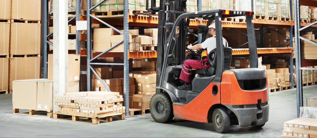 A warehouse scene with a worker operating an orange forklift among aisles of neatly stacked cardboard boxes on metal shelving units. The scene captures a moment of everyday logistics and inventory management in a distribution center.