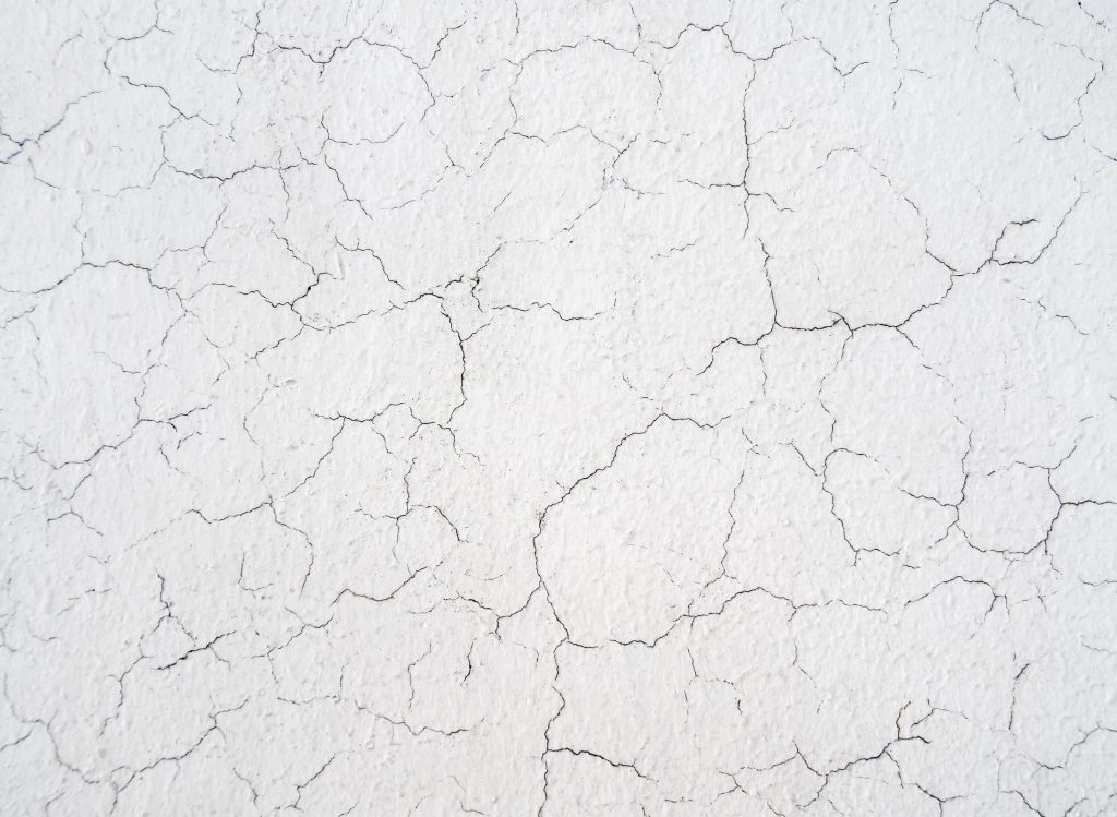 A close-up of a white wall with a cracked paint texture, showing a network of fine lines and splits that create an intricate pattern of distress on the surface called crazing.