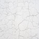 A close-up of a white wall with a cracked paint texture, showing a network of fine lines and splits that create an intricate pattern of distress on the surface called crazing.