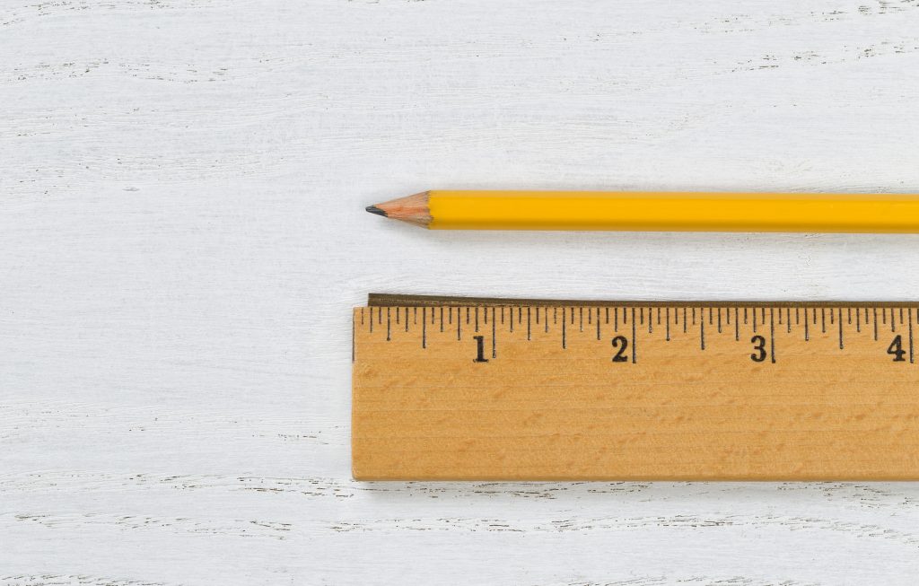 A classic yellow pencil and a wooden ruler displayed horizontally on a white distressed wood surface. The ruler shows measurements in inches, and the sharp pencil tip points towards the left, indicating readiness for writing or drawing.