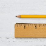 A classic yellow pencil and a wooden ruler displayed horizontally on a white distressed wood surface. The ruler shows measurements in inches, and the sharp pencil tip points towards the left, indicating readiness for writing or drawing.