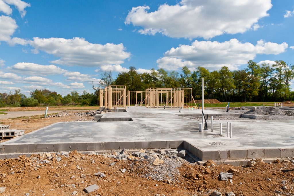 Early construction phase of a new building with the new concrete foundation laid out and partial wooden framing erected. Plumbing stubs are visible on the concrete slab, and there is a clear sky above the construction site, which is surrounded by a natural landscape. The image depicts the beginnings of development in a spacious outdoor setting.