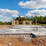 Early construction phase of a new building with the new concrete foundation laid out and partial wooden framing erected. Plumbing stubs are visible on the concrete slab, and there is a clear sky above the construction site, which is surrounded by a natural landscape. The image depicts the beginnings of development in a spacious outdoor setting.