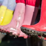 Close-up of three pairs of colorful rubber boots—red, yellow, and pink—covered in mud. The boots are hanging over the edge, likely on a bench, with their soles showing dirt from recent use. The focus on the boots conveys an outdoor, adventurous setting, possibly after a day of gardening or walking in the rain.