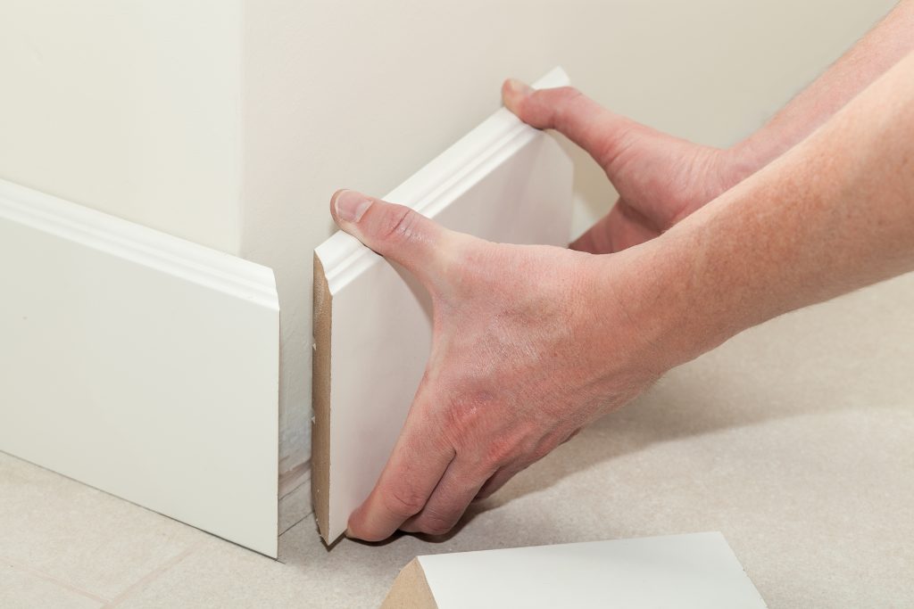 Close-up of hands installing white baseboard molding on a wall, with precise alignment at the corner joint. The person is pressing the two adjoining pieces together to ensure a snug fit, illustrating a step in interior finish carpentry.