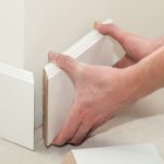 Close-up of hands installing white baseboard molding on a wall, with precise alignment at the corner joint. The person is pressing the two adjoining pieces together to ensure a snug fit, illustrating a step in interior finish carpentry.