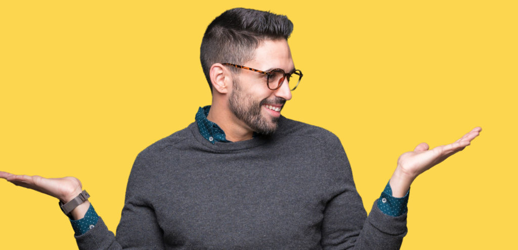 A cheerful man with a beard and glasses, wearing a grey sweater over a collared shirt, playfully shrugs with his palms facing up against a bright yellow background suggesting options of color flakes or color granules in epoxy flooring.