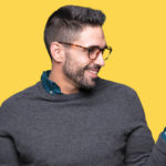 A cheerful man with a beard and glasses, wearing a grey sweater over a collared shirt, playfully shrugs with his palms facing up against a bright yellow background suggesting options of color flakes or color granules in epoxy flooring.