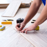 A person in blue attire is installing laminate flooring, measuring and marking the wooden plank to fit. Flooring installation tools, such as a handsaw, clamps, a tape measure, and other equipment, are scattered around the work area. The focus is on the hands performing a precise measurement, indicating skilled manual work.