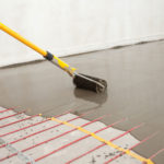 Application of a self-leveling compound on a concrete floor using a spiked roller attached to a yellow pole. The floor has underfloor heating pipes laid out in a grid pattern, visible through the semi-liquid layer of the compound. This image represents a stage in preparing a floor for final surface installation.