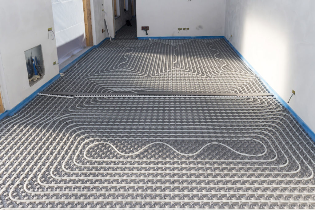A radiant floor heating system installation in progress, showing a network of interconnected pipes laid out in a serpentine pattern across a room. The floor is prepared with insulation panels underneath the pipes, and walls are primed for finishing, with electrical outlets and wiring visible. The image illustrates a modern method of efficient home heating.