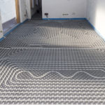 A radiant floor heating system installation in progress, showing a network of interconnected pipes laid out in a serpentine pattern across a room. The floor is prepared with insulation panels underneath the pipes, and walls are primed for finishing, with electrical outlets and wiring visible. The image illustrates a modern method of efficient home heating.