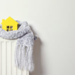 A simplistic yellow paper house perched on a white radiator, wrapped in a cozy grey knitted scarf with tassels. The scene evokes a sense of warmth and home comfort, suggesting energy-saving or staying warm during winter.