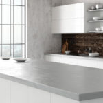 A modern kitchen interior with a large window providing a cityscape view. The kitchen features a minimalist design with a white concrete countertop, bar stools, and a wooden backsplash. Open shelving holds neatly arranged dishes and kitchenware. Natural light fills the room, creating a bright and airy atmosphere.