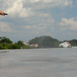 A worker in a blue uniform and rubber boots using a hose to clean a large concrete surface on a rooftop, with a village and cloudy sky in the background.