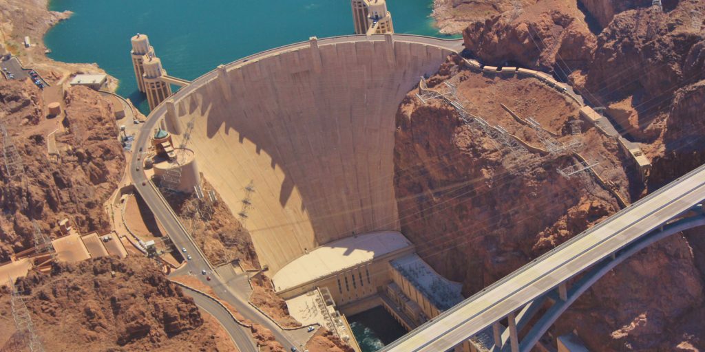 Aerial view of the Hoover Dam, showcasing the massive concrete arch-gravity structure, the power plant buildings at its base, and the Mike O'Callaghan–Pat Tillman Memorial Bridge in the background, set amidst the rugged landscape.