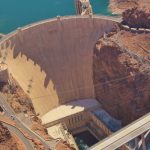 Aerial view of the Hoover Dam, showcasing the massive concrete arch-gravity structure, the power plant buildings at its base, and the Mike O'Callaghan–Pat Tillman Memorial Bridge in the background, set amidst the rugged landscape.