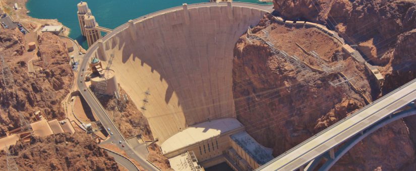 Aerial view of the Hoover Dam, showcasing the massive concrete arch-gravity structure, the power plant buildings at its base, and the Mike O'Callaghan–Pat Tillman Memorial Bridge in the background, set amidst the rugged landscape.