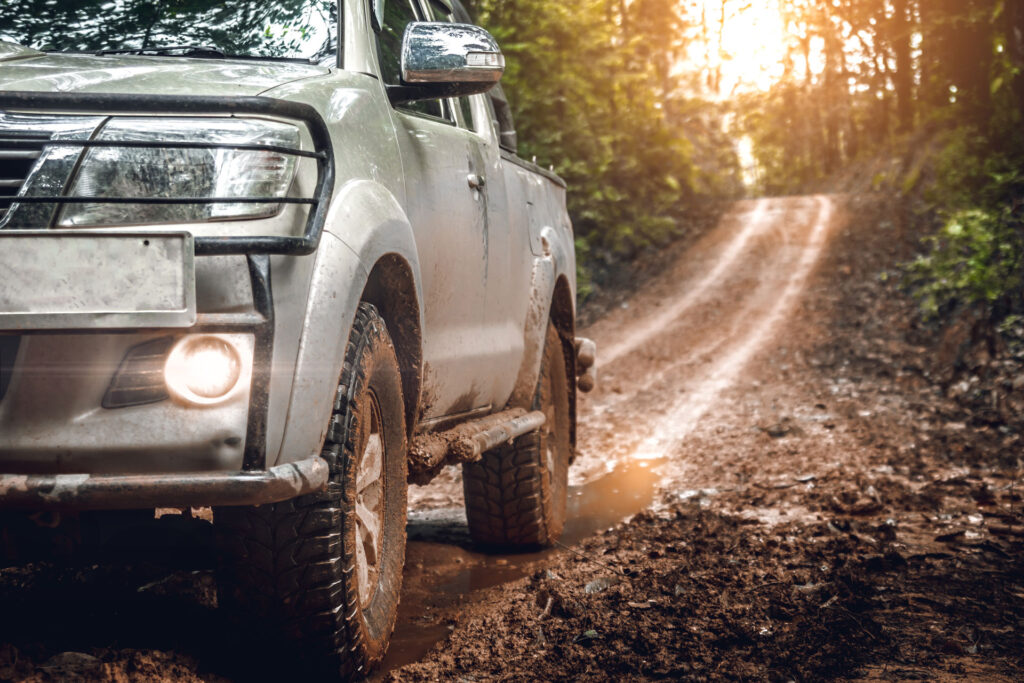 A close-up of a muddy off-road vehicle's front wheel and partial view of the grille, with headlights on, set against a backdrop of a forest trail illuminated by sunlight.