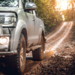 A close-up of a muddy off-road vehicle's front wheel and partial view of the grille, with headlights on, set against a backdrop of a forest trail illuminated by sunlight.