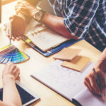 A team collaborating around a table with design tools: a person holding a color swatch, another drawing a flowchart, and samples of wood veneer alongside a laptop and tablet, indicative of a creative brainstorming session.