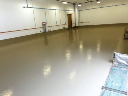 A spacious room with newly applied shiny beige epoxy flooring reflecting ceiling lights, surrounded by white walls with blue and copper piping, indicating an industrial or commercial setting.