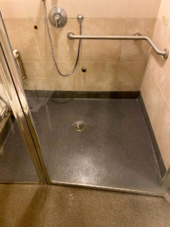 Accessible shower area with dark brown non-slip floor, equipped with stainless steel grab bars, a flexible shower hose, and a clear glass door, set against light brown tiled walls.