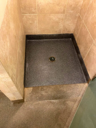 Corner shower area with textured dark brown epoxy floor and light brown tiled walls, featuring a central drain with a black and white mosaic cover.