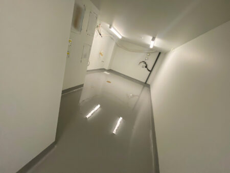 An interior view of a room under construction with freshly applied grey epoxy flooring that reflects the ceiling lights, surrounded by white walls with exposed electrical wiring and fixtures.