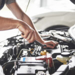 A mechanic's hands using a wrench on a car engine, showcasing a close-up of automotive maintenance work with various engine components and a car battery in the foreground.