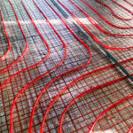 Close-up of a red underfloor heating installation with winding pipes laid out on a mesh support over a concrete substrate, illustrating the process of installing a radiant floor heating system.